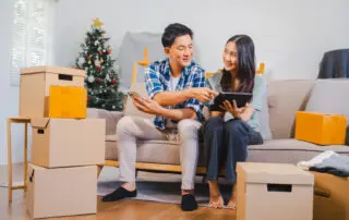 Couple is unpacking boxes in a festive living room with a Christmas tree, showing surprise and happiness with a computer checklist