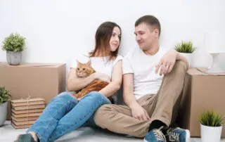 A young married couple with a red cat, a man and a woman, are sitting on the floor in a bright room against the background of cardboard boxes, green plants and things.