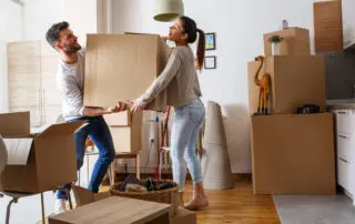 Man and woman carrying a moving box through a room with other moving boxes.