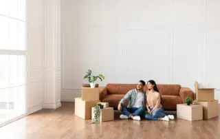 Moving. Cheerful Spouses Sitting Among Unpacked Moving Boxes In New Home, Hugging And Looking Aside At Window.