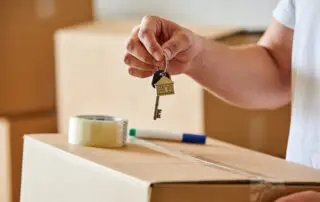 Close-up of hands holding keys symbolizing new home ownership, with blurred boxes in the background. A warm atmosphere highlighting real estate transition.
