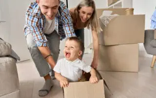 Happy parents having fun playing with cute toddler kid son riding in box on moving day. Excited young couple pushing box with child boy celebrating relocation. Young family move into new home