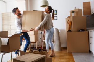 Man and woman carrying a moving box through a room with other moving boxes.