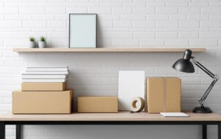 Home office setting featuring cardboard boxes, books, stacked lamp near minimalist shelving against white brick wall, suggesting small business workspace