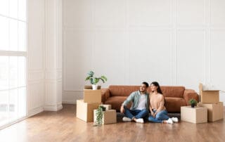 Moving. Cheerful Spouses Sitting Among Unpacked Moving Boxes In New Home, Hugging And Looking Aside At Window.
