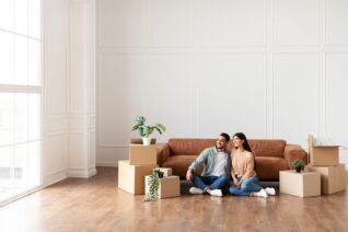 Moving. Cheerful Spouses Sitting Among Unpacked Moving Boxes In New Home, Hugging And Looking Aside At Window.