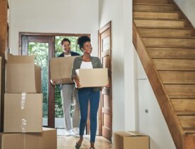 couple moving into a new modern house, carrying boxes and arriving home together. Happy, excited and smiling husband and wife walking, entering and relocating.