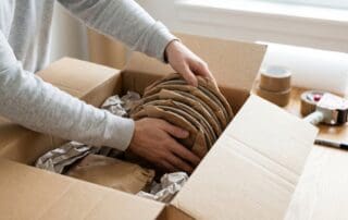 Person carefully wrapping ceramic plates in brown packing paper and placing them in a moving box