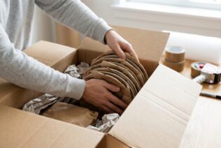 Person carefully wrapping ceramic plates in brown packing paper and placing them in a moving box
