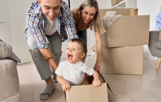 Happy parents having fun playing with cute toddler kid son riding in box on moving day. Excited young couple pushing box with child boy celebrating relocation. Young family move into new home