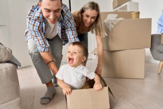 Happy parents having fun playing with cute toddler kid son riding in box on moving day. Excited young couple pushing box with child boy celebrating relocation. Young family move into new home
