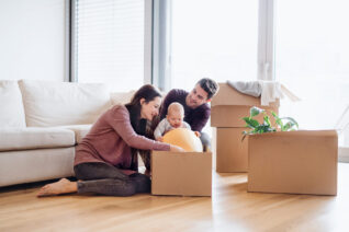 A happy young couple sitting on the floor with a baby and cardboard boxes, moving in a new home.